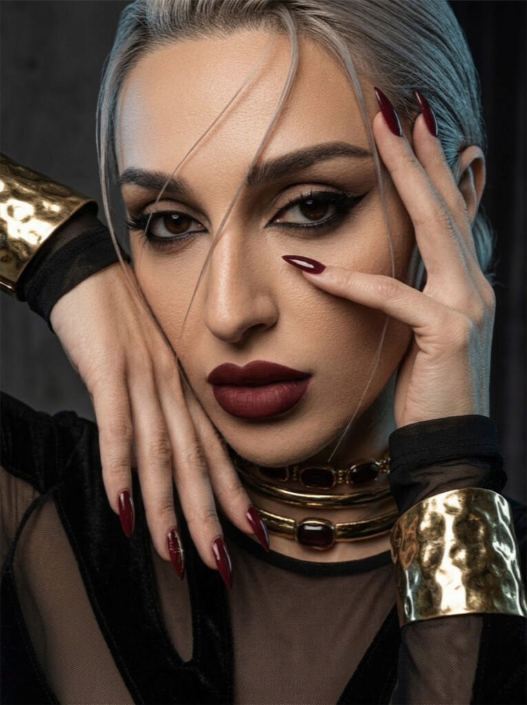 Close-up portrait of Aylin Baykal wearing layered gold Starchuska necklaces and a wide cuff, with dark red nails and makeup.