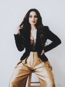Aylin Baykal seated on a stool in a black blazer and gold wide-leg trousers for Starchuska Women’s AW’26.