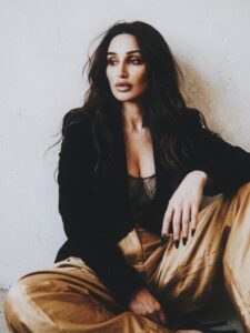 Aylin Baykal seated against a white wall in a black blazer and gold trousers for Starchuska Women’s AW’26.