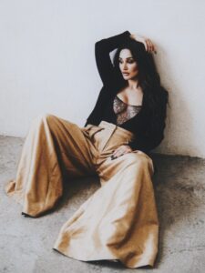 Aylin Baykal reclining against a white wall in black tailoring and wide-leg gold trousers.