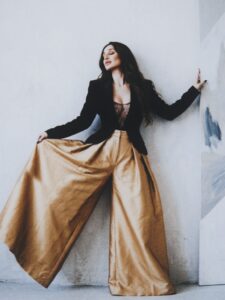 Aylin Baykal posing against a white studio wall in a black blazer and flowing gold trousers.