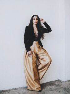 Aylin Baykal posing with one leg raised in a black blazer and gold trousers for Starchuska Women’s AW’26.