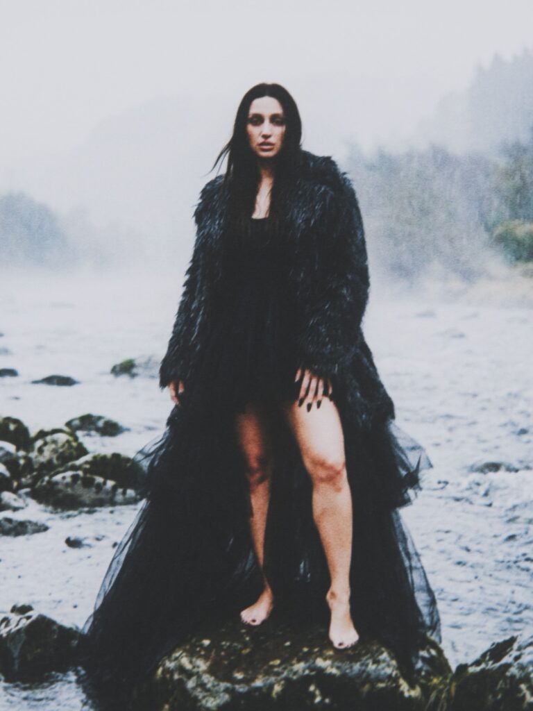 Aylin Baykal barefoot on a rock in a black textured coat and flowing sheer dress for Starchuska Women’s AW’26.