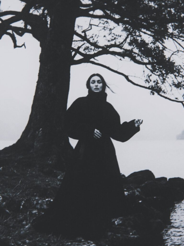 Black-and-white portrait of Aylin Baykal in a long dark coat beneath a tree by the water for Starchuska Women’s AW’26.