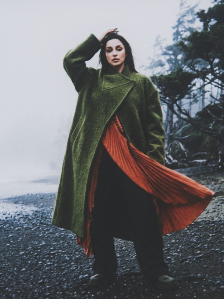 Aylin Baykal wearing a sculptural green coat over a rust pleated dress on a rocky shoreline for Starchuska Women’s AW’26.