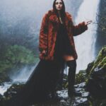 Aylin Baykal in a rust faux-fur coat and black dress beside a misty waterfall for Starchuska Women’s AW’26.