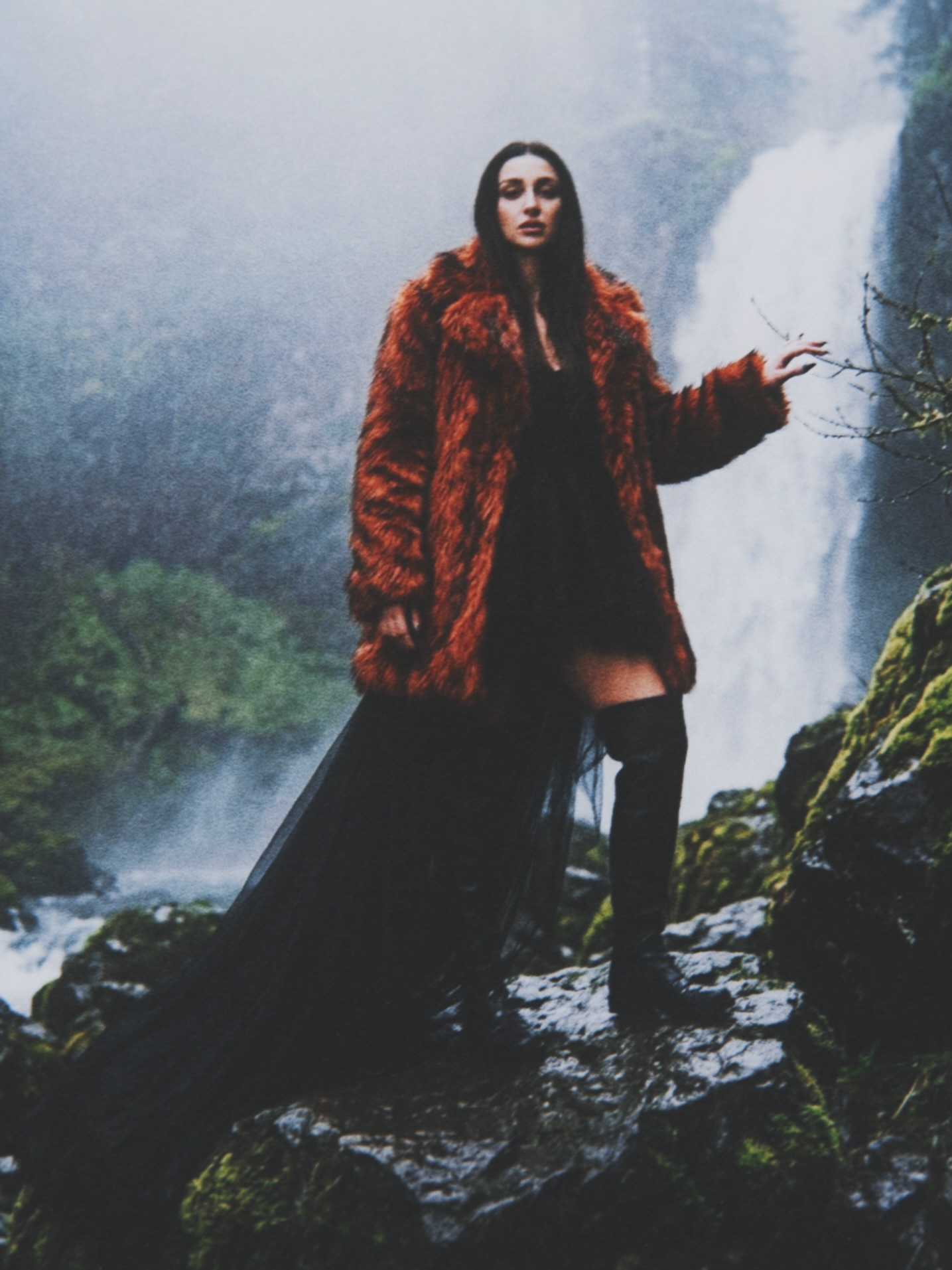 Aylin Baykal in a rust faux-fur coat and black dress beside a misty waterfall for Starchuska Women’s AW’26.